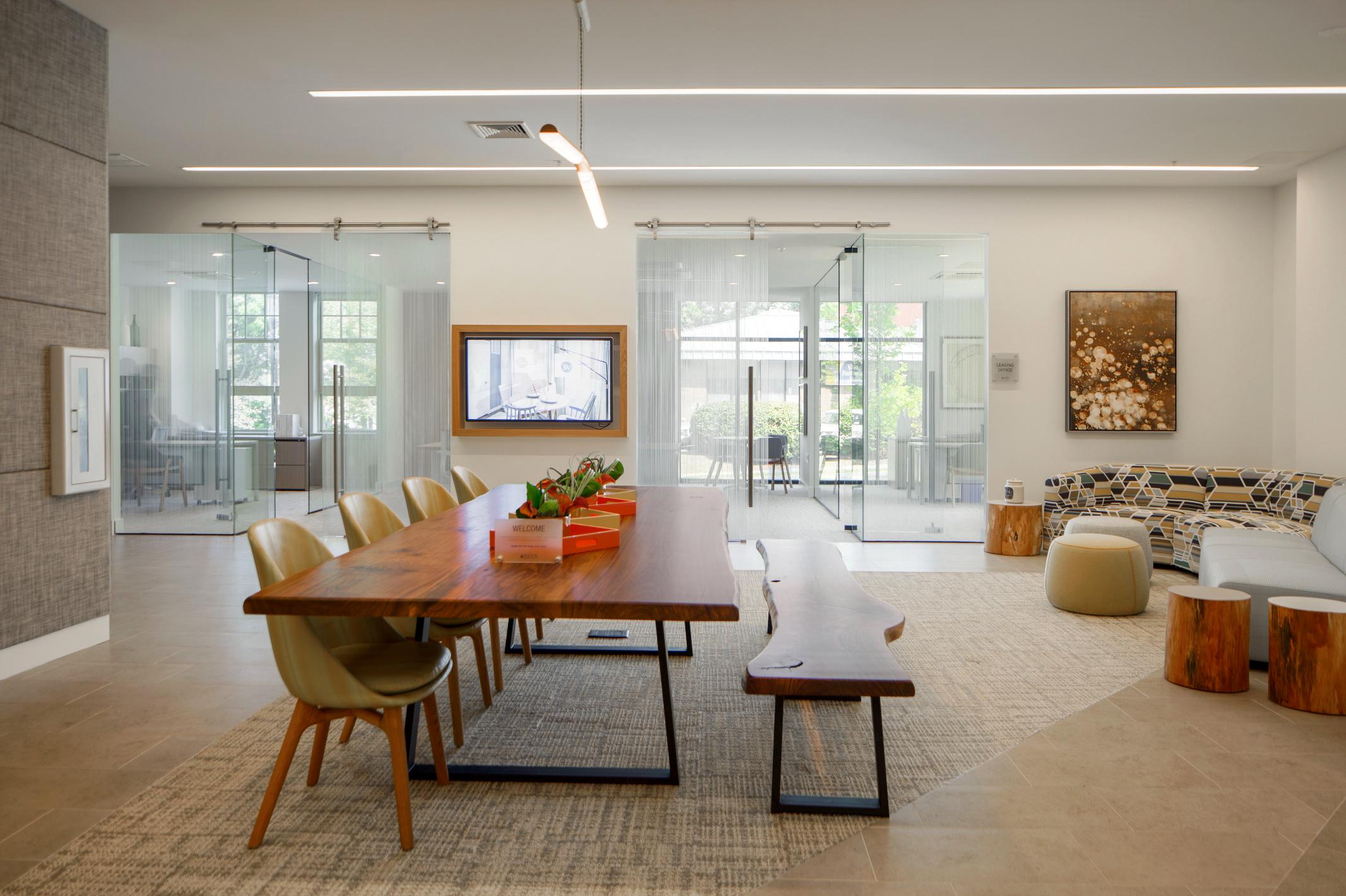 A long wooden table with chairs and a bench in a modern lounge, with glass-walled offices