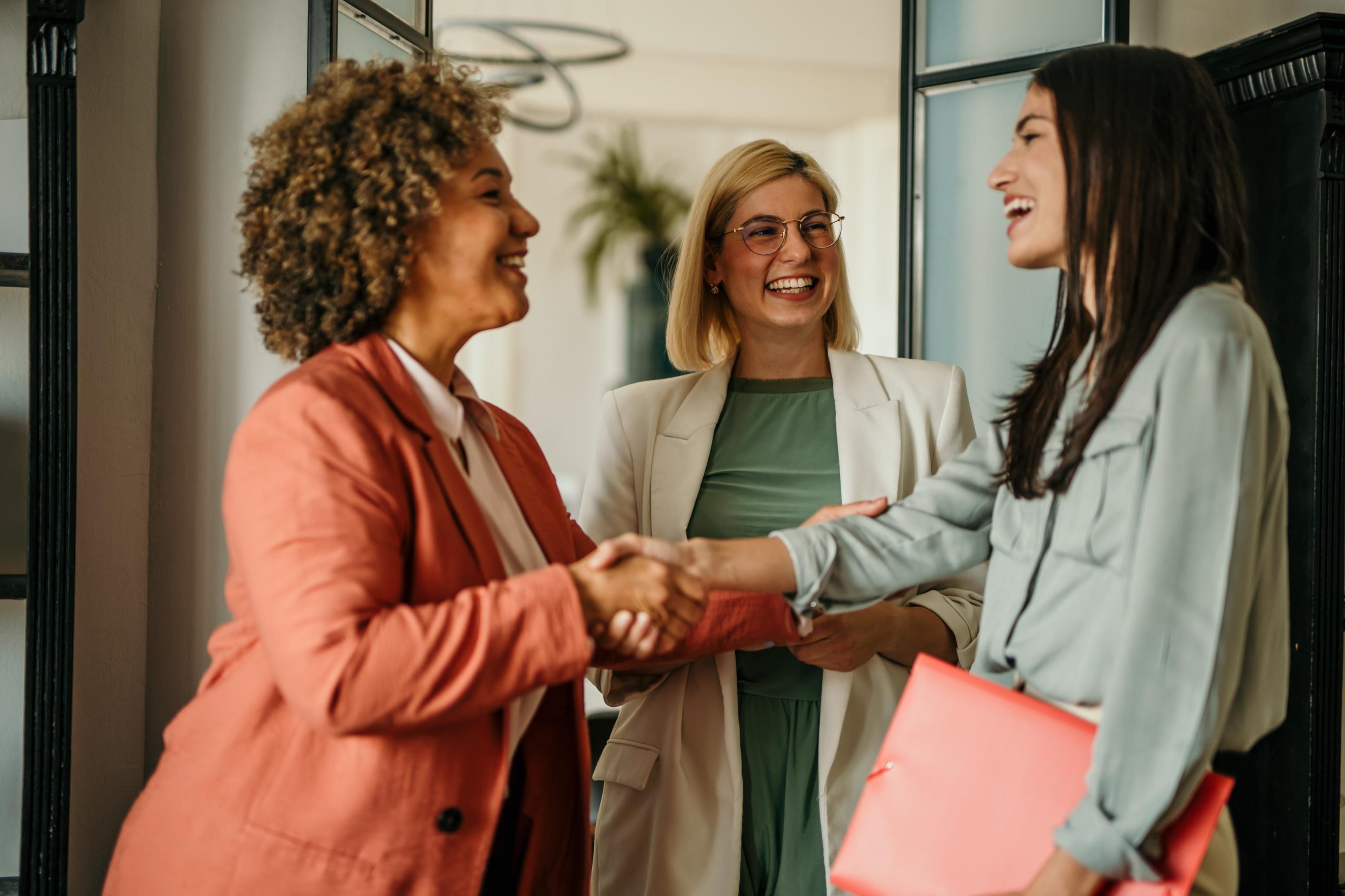 A group of three professional women in suits are smiling and shaking hands.