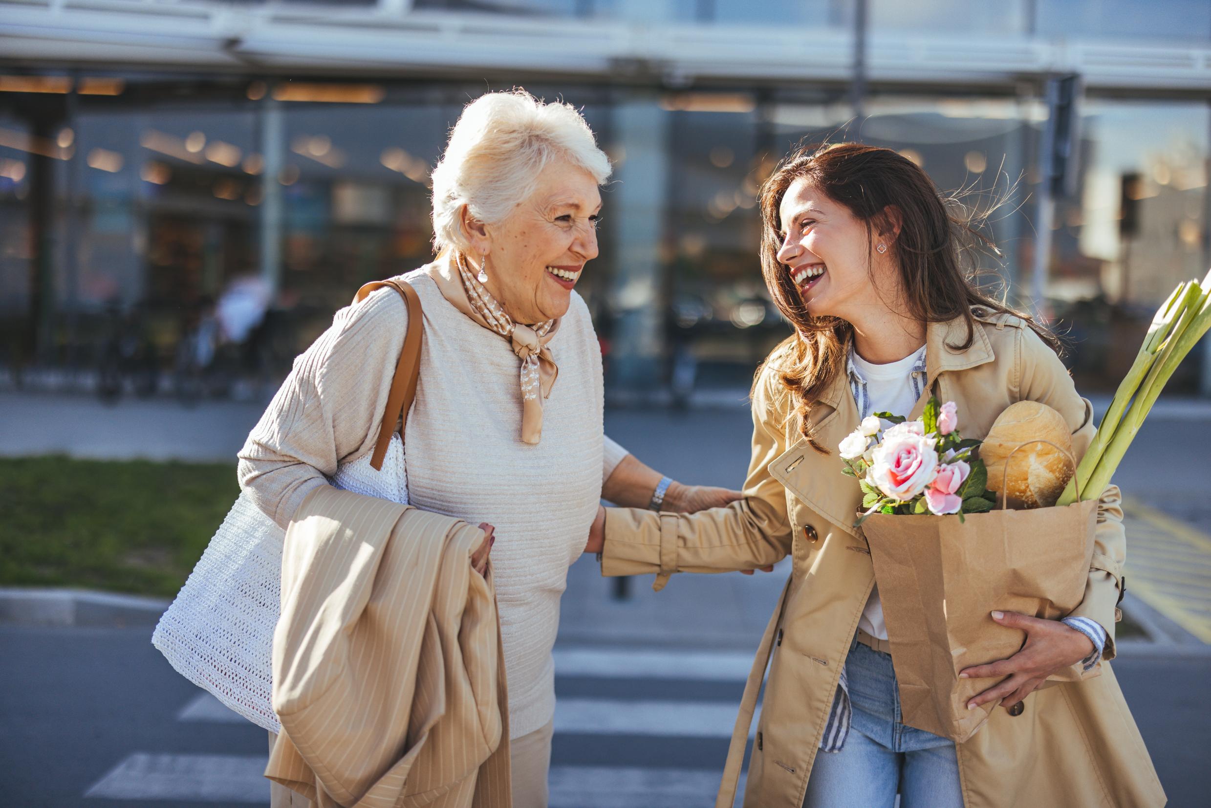 An elderly and a middle-aged woman are crossing the street while holding groceries and smiling.